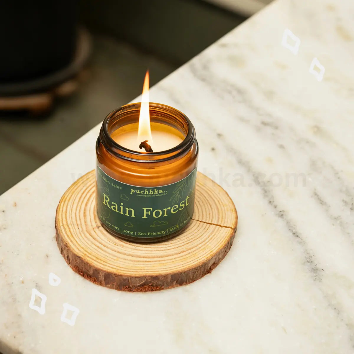 A lit amber glass jar candle labeled 'Rain Forest' by Puchhka, resting on a round wooden coaster on a white marble surface.