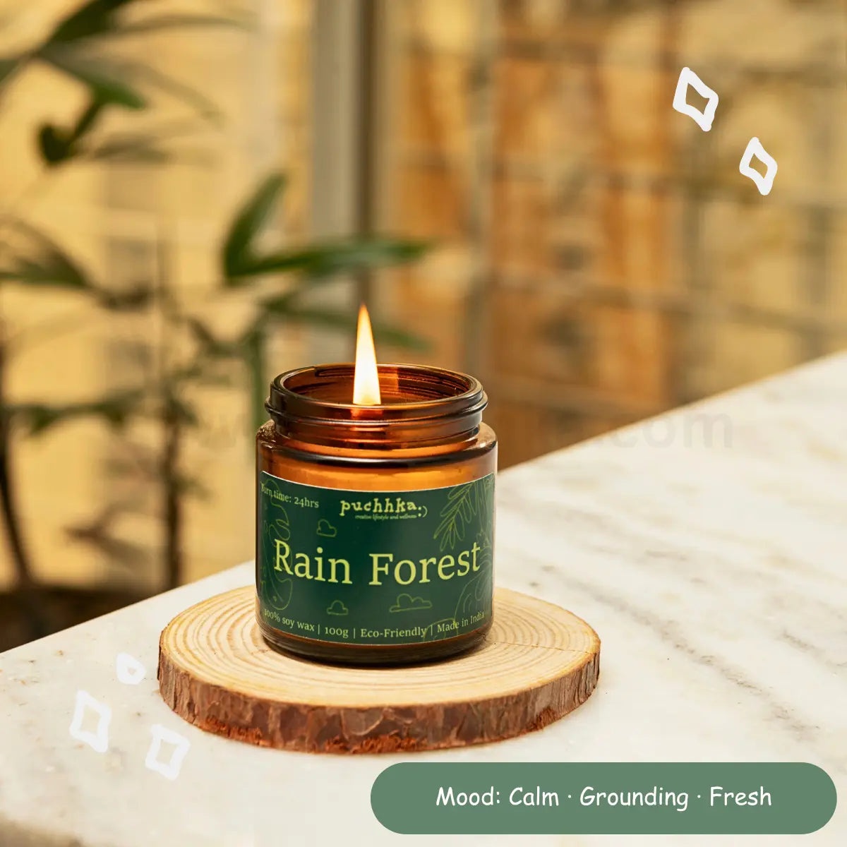 A lit amber glass candle jar labeled 'Rain Forest' by Puchhka, resting on a round wooden coaster on a marble surface with a blurred indoor background.