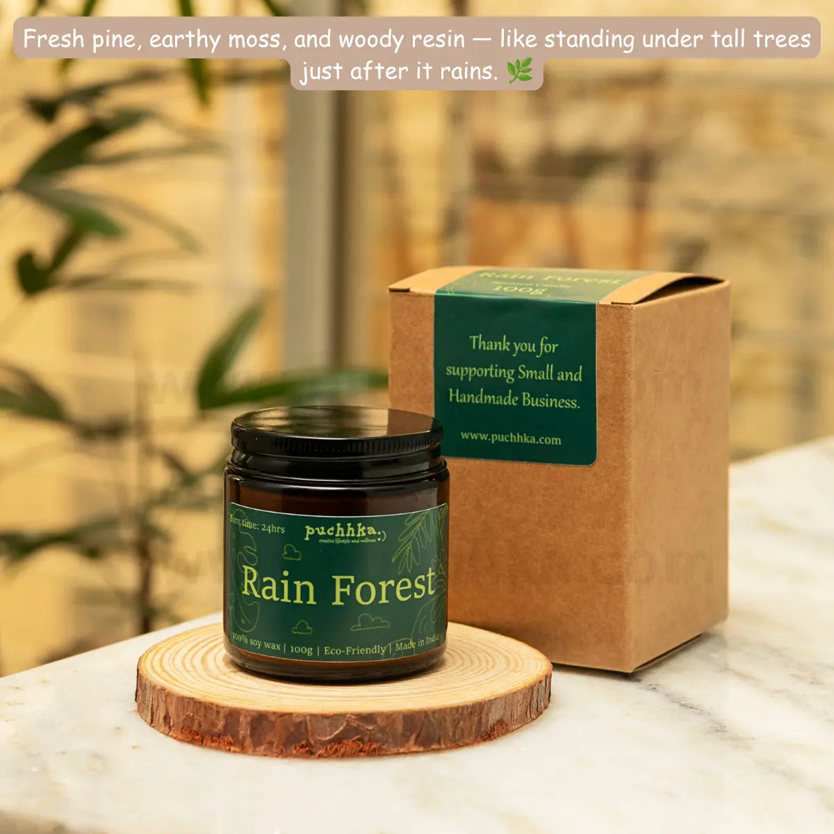A jar of Puchhka 'Rain Forest' scented candle sits on a round wooden coaster next to its kraft paper gift box, set on a white marble countertop with a blurred indoor background.