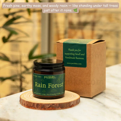 A jar of Puchhka 'Rain Forest' scented candle sits on a round wooden coaster next to its kraft paper gift box, set on a white marble countertop with a blurred indoor background.