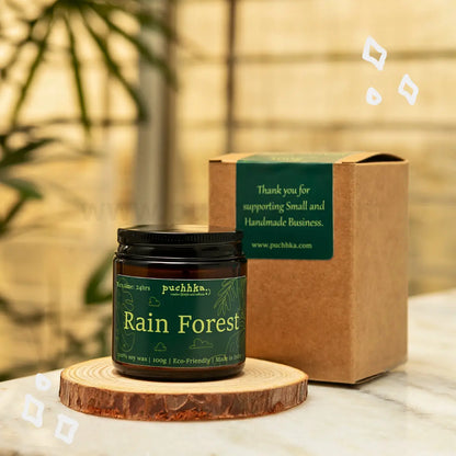 A jar of Puchhka Rain Forest scented candle stands on a round wooden coaster next to its brown kraft paper box, set on a marble surface with a warm, blurred background.