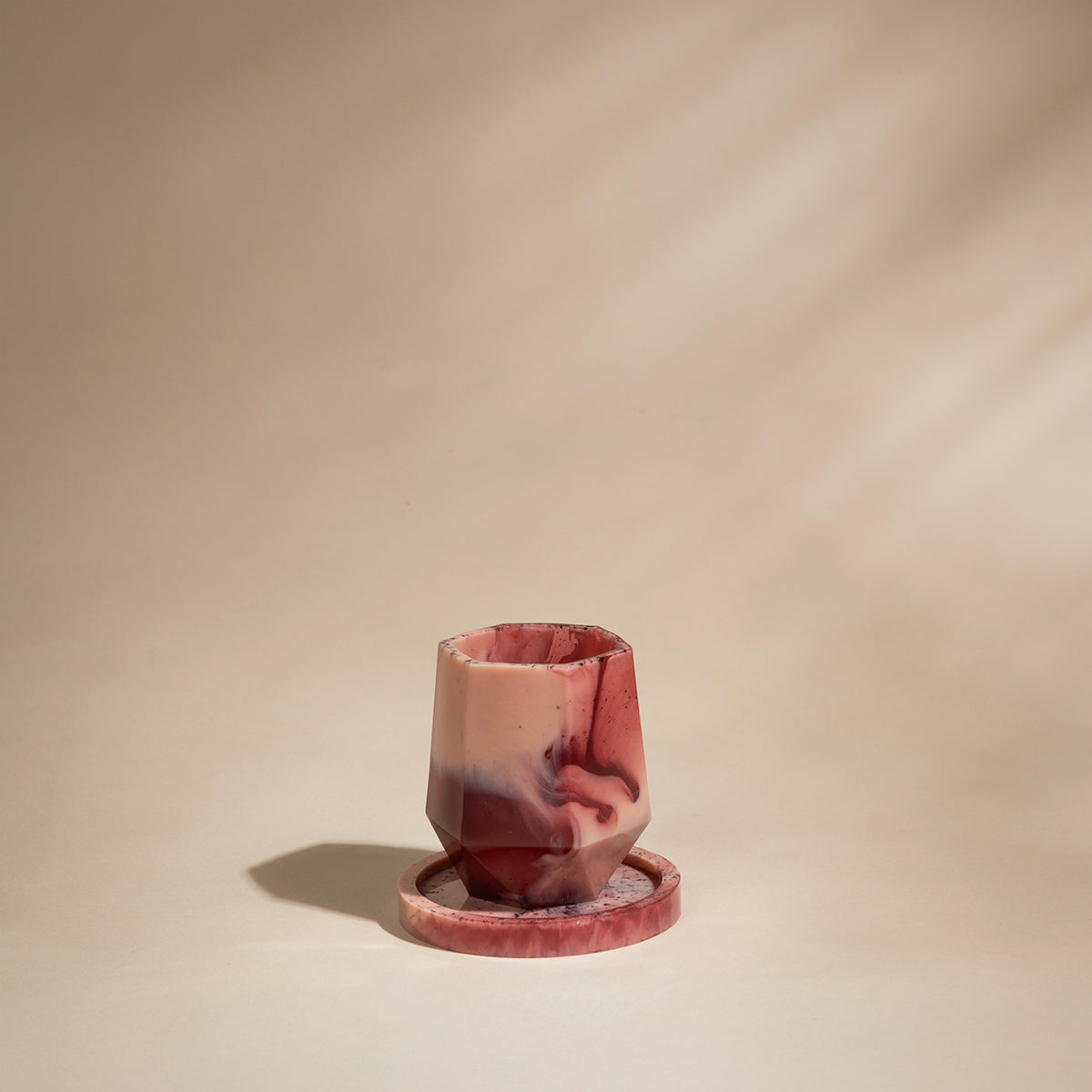 A faceted resin tumbler with a matching coaster, both made from a resin in shades of pink, red, and cream, set against a warm beige background with soft diagonal shadows.