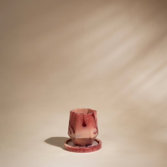 A small sculptural tumbler made of faceted translucent pink and burgundy resin, shown on a matching round base against a warm beige studio background with soft diagonal shadows.