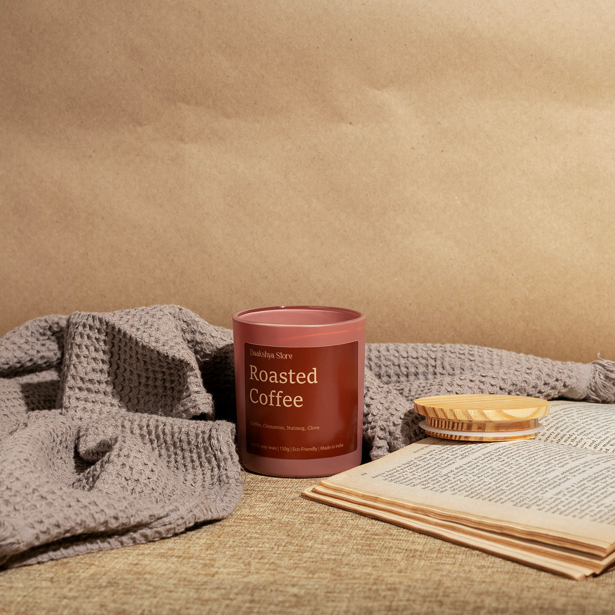 A candle jar labeled 'Roasted Coffee' by Daakshya Store, styled on a woven surface with a gray knit blanket, an open book, and a small wooden dish against a tan paper backdrop.