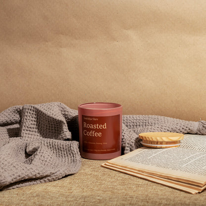 A candle jar labeled 'Roasted Coffee' by Daakshya Store, styled on a woven surface with a gray knit blanket, an open book, and a small wooden dish against a tan paper backdrop.