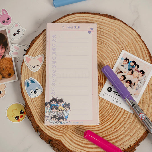 A pink notepad with a checklist design and cartoon character illustration at the bottom, styled on a round wood slice with stickers, a purple pen, a pink marker, and a printed photo on a light marble surface.