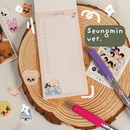 A pastel pink checklist notepad with cute cartoon characters and the text '5 o'clock List' is displayed on a rustic wood slice. The notepad is styled in a flat lay with a purple and pink pen, a photo print, and various animal stickers, with a green text bubble reading 'Seungmin ver.