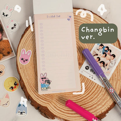 A pastel pink notepad with lined writing space and cute cartoon bunny illustrations, displayed on a round wood slice with pens, stickers, and a printed photo around it. A dark green label reads 'Changbin ver.'