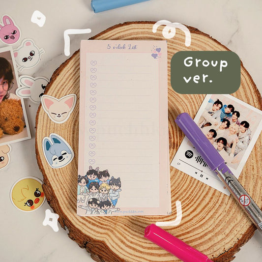 A pink notepad with a checklist and cartoon character illustration at the bottom, styled on a round wood slice with stickers, a purple marker, a pink marker, and group photo prints. A green label reads 'Group ver.