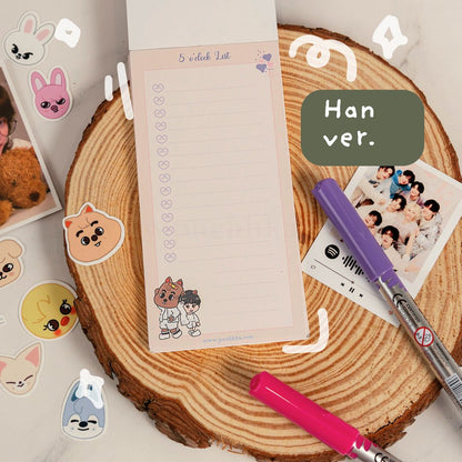 A cute K-pop themed stationery set featuring a checklist notepad with a cartoon cat and boy illustration, displayed on a rustic wood slice with colorful pens, stickers, and a photo print.