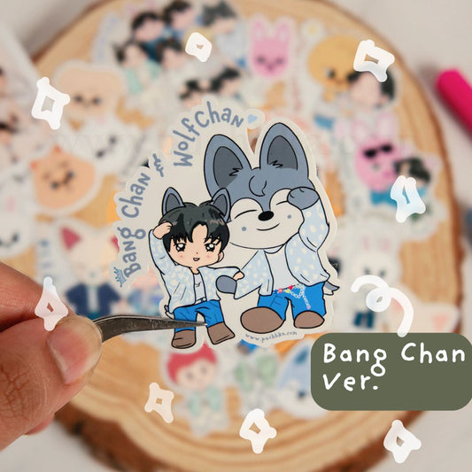 A hand holds a die-cut sticker featuring chibi character labeled 'Bang Chan' and 'Wolf Chan' on a wooden slice background with other stickers blurred around it.