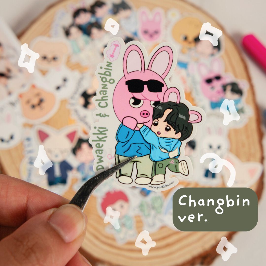 A hand holds a die-cut sticker featuring a pink bunny character wearing sunglasses and hugging a person with black hair, with the text 'Dwaekki & Changbin' and 'Changbin ver.' on the sticker.