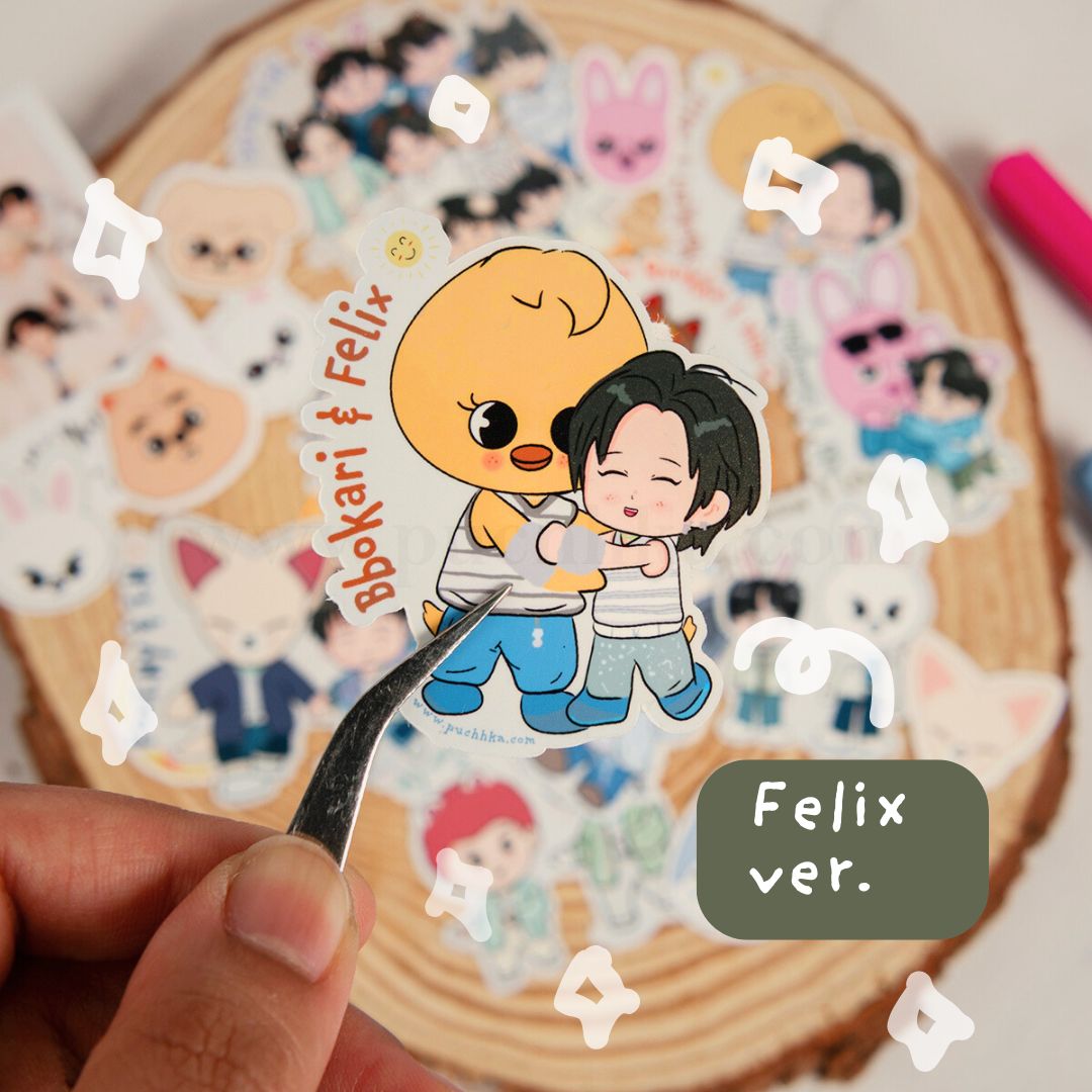 A hand holds a die-cut sticker featuring two cartoon characters hugging, with the text 'Bbokari & Felix' and 'Felix ver.' over a round wooden display filled with other stickers.