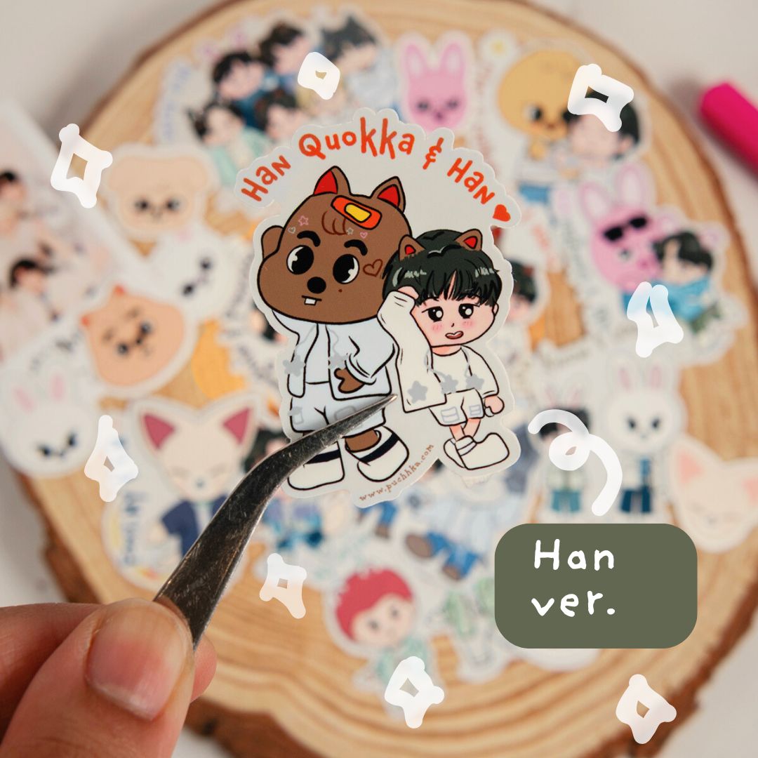 A close-up of a hand holding tweezers with a small die-cut sticker featuring two cartoon characters and the text 'Han Quokka & Han ver.' over a stack of other stickers on a wood slice background.