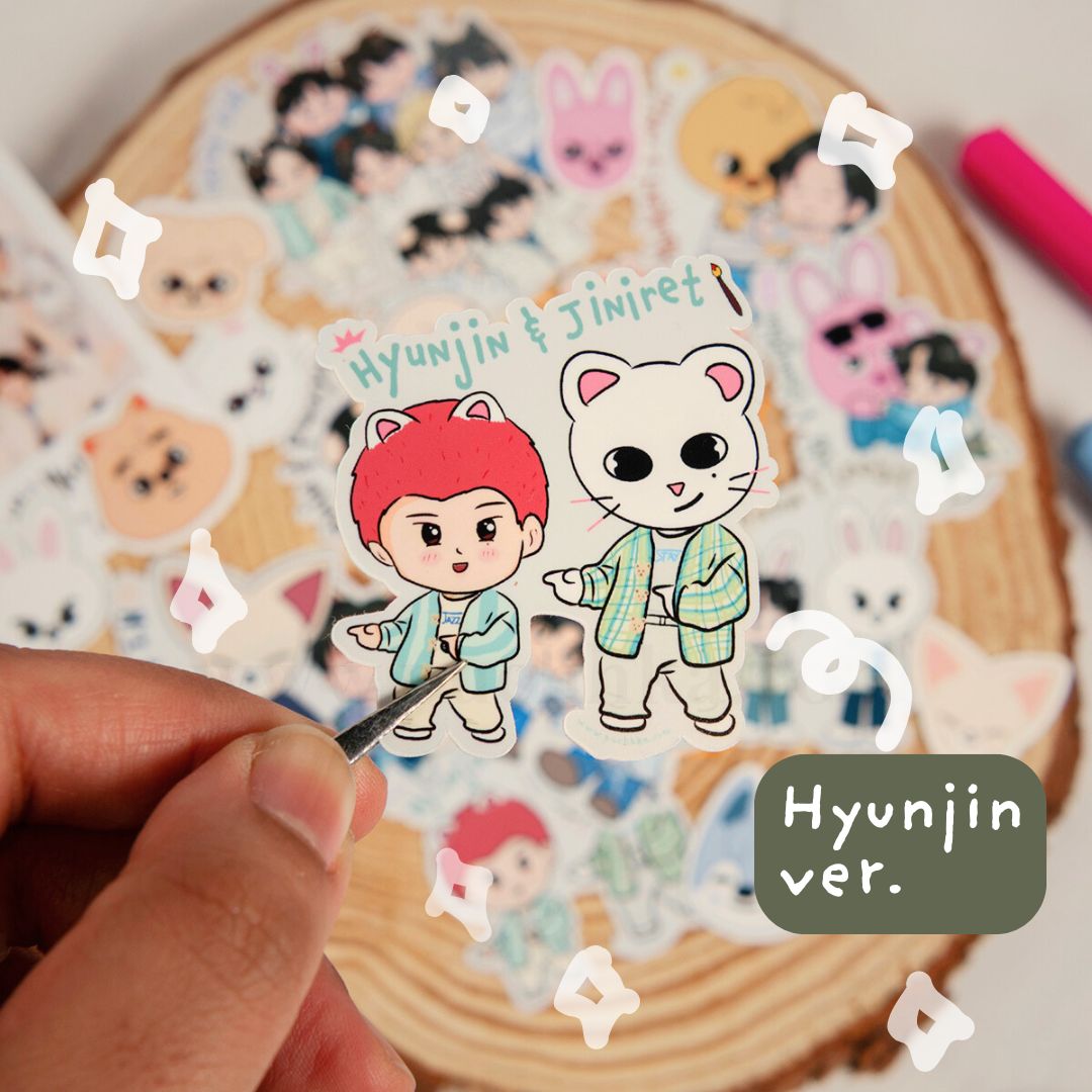 A hand holds a die-cut sticker featuring two chibi characters labeled 'Hyunjin & Jiniret' and 'Hyunjin ver.' against a background of many other colorful stickers on a wooden slice.