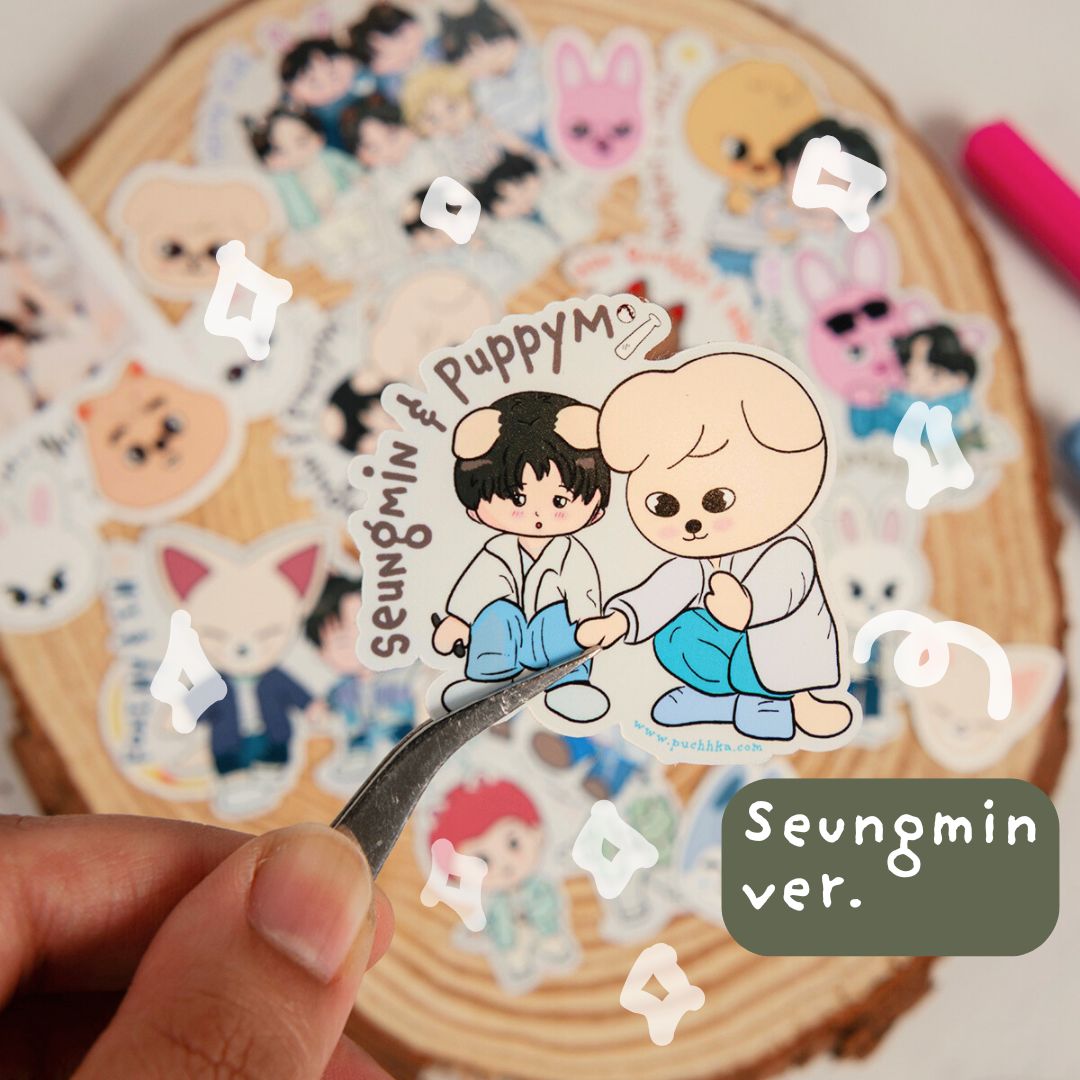 A hand holds a die-cut sticker featuring two cartoon characters with the text 'Seungmin & Puppym Seungmin ver.' over a stack of similar stickers on a wood slice background.
