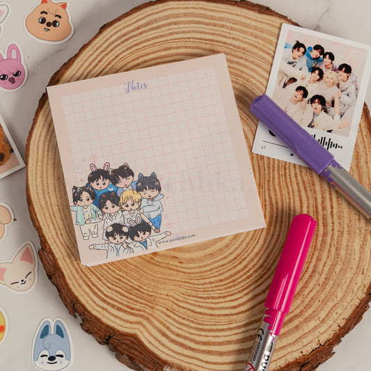 A square grid notepad with chibi stray kids group illustration and the word 'Notes' at the top, styled on a round wood slice with pens and a printed photo nearby.
