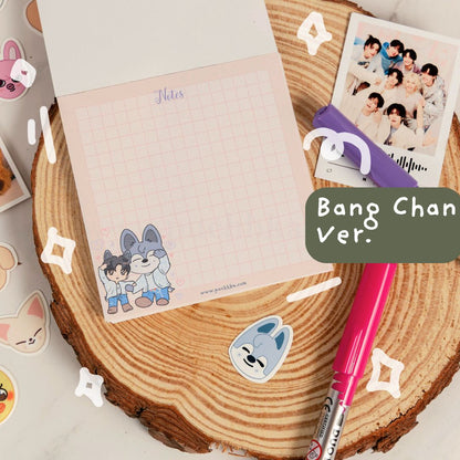 A pink grid notepad with skz illustrations, styled on a wooden coaster with a pink pen, purple highlighter, instant photo, and stickers. A green label reads 'Bang Chan Ver.' 