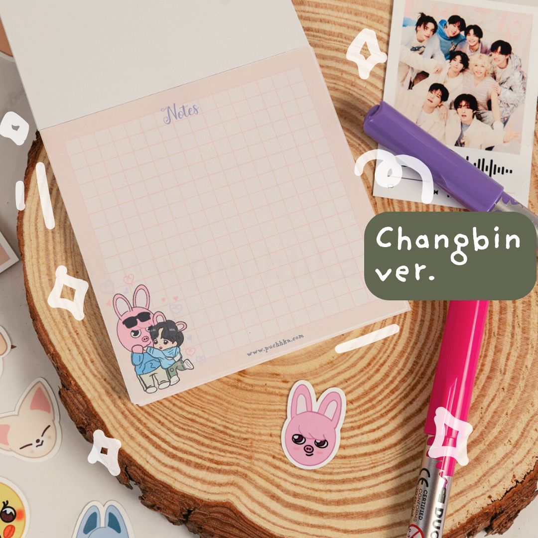 A pastel pink grid notepad with a cute dwaekki illustration and the text 'Notes' at the top, styled on a wood slice with a purple marker, pink pen, sticker, and a photo print. A green label reads 'Changbin ver.