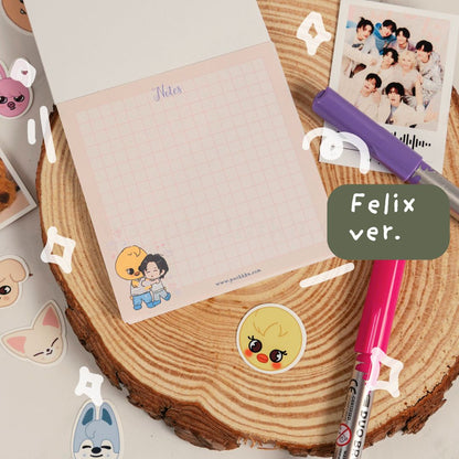 A pastel pink grid notepad with a cute chibi sticker on the bottom left, styled on a round wood slice with pens, stickers, and a printed photo. A green label reads 'Felix ver.