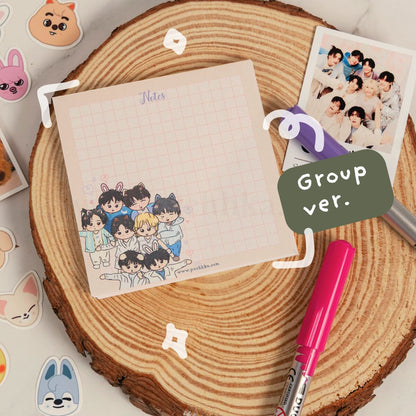 A cute group illustration notepad labeled 'Notes' and 'Group ver.' displayed on a round wood slice with a pink marker, a group photo, and decorative stickers around it.