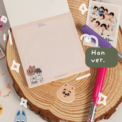 A pink grid notepad with chibi han  stickers displayed on a round wood slice, styled with a pink pen, a purple highlighter, a printed photo, and decorative sticker elements.