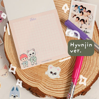 A styled flat-lay of a pink grid notepad with cute chibi hyunjin stickers, placed on a wood slice background with pens, stickers, and a printed photo. A green label reads 'Hyunjin ver.