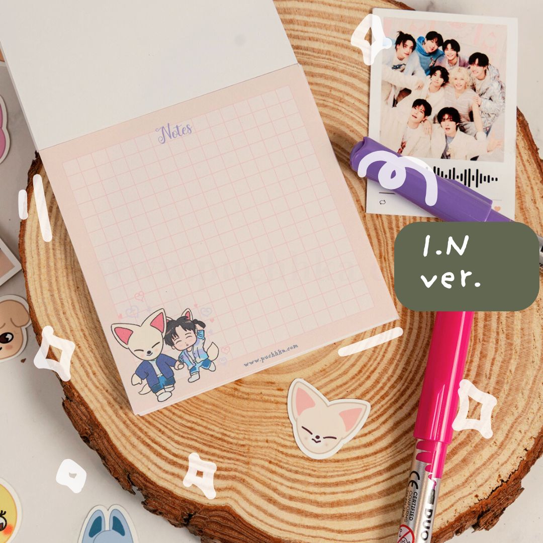 A pastel pink grid notepad with cute skz illustrations at the bottom, styled on a round wood slice with a pink pen, a purple marker, a printed photo, and decorative stickers. A dark green label reads 'I.N'.
