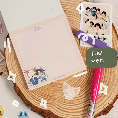 A pastel pink grid notepad with cute skz illustrations at the bottom, styled on a round wood slice with a pink pen, a purple marker, a printed photo, and decorative stickers. A dark green label reads 'I.N'.