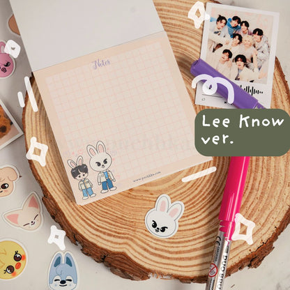 A pastel grid notepad with chibi stray kids illustrations, shown on a wood slice with a pink pen, a printed photo, stickers, and a green label reading 'Lee Know ver.'.