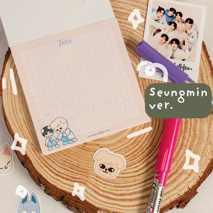 A pink grid notepad with skz illustrations and the text 'Notes' and 'Seungmin ver.' is displayed on a rustic wood slice background, styled with a pink pen, purple pen, and a printed photo of a group of boys.