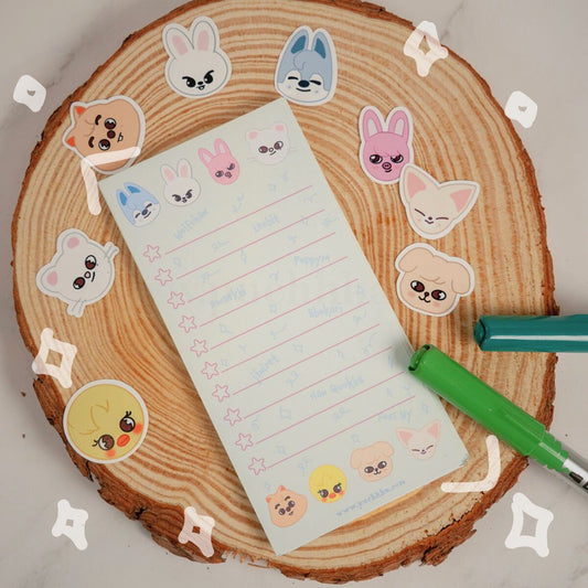 A cute notepad with skzoos arranged on a round wood slice, with two pens nearby on a light marble surface.