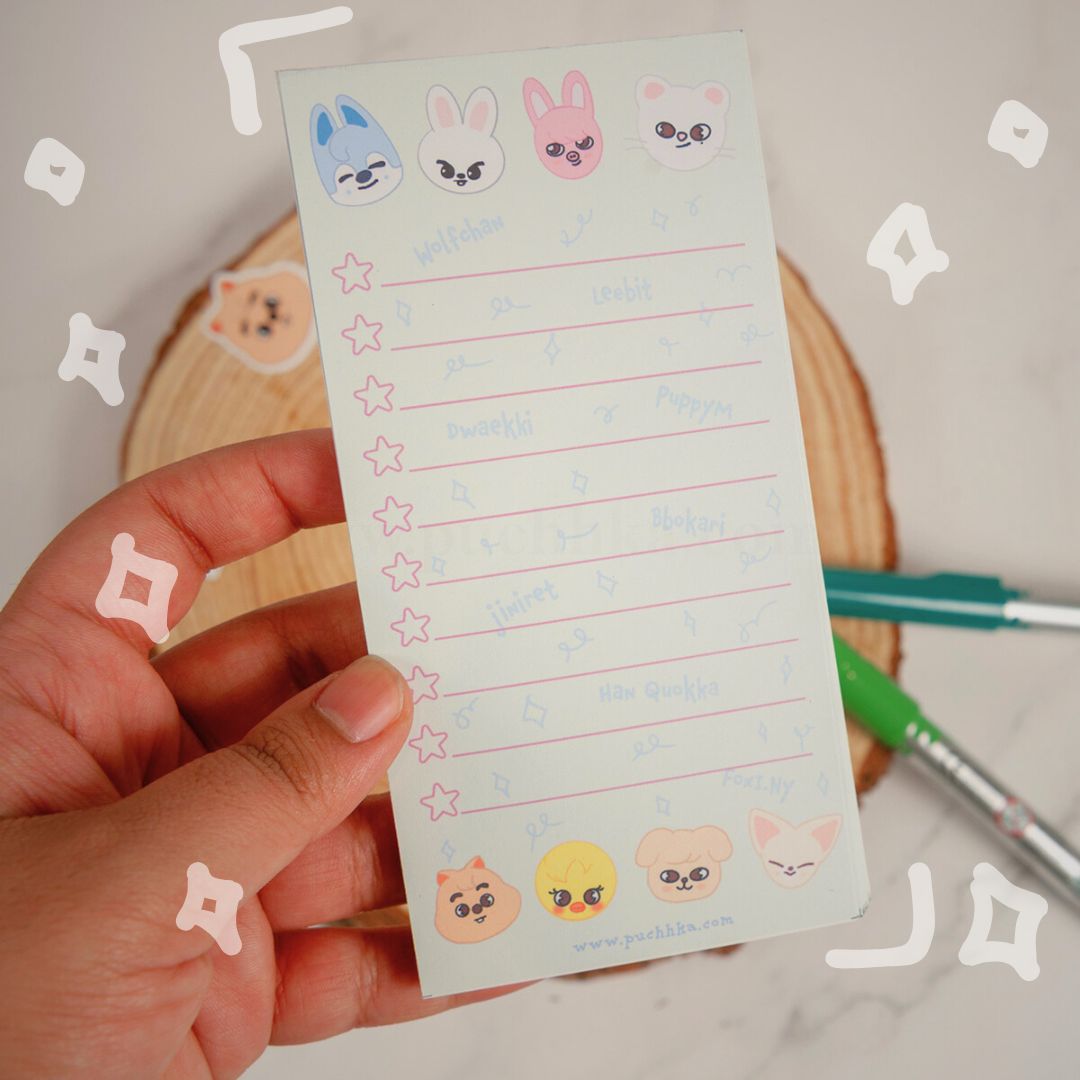 A hand holds a small notepad with cute skzoo faces and lined writing sections.  The photo is styled with a wooden coaster and pens in the background.