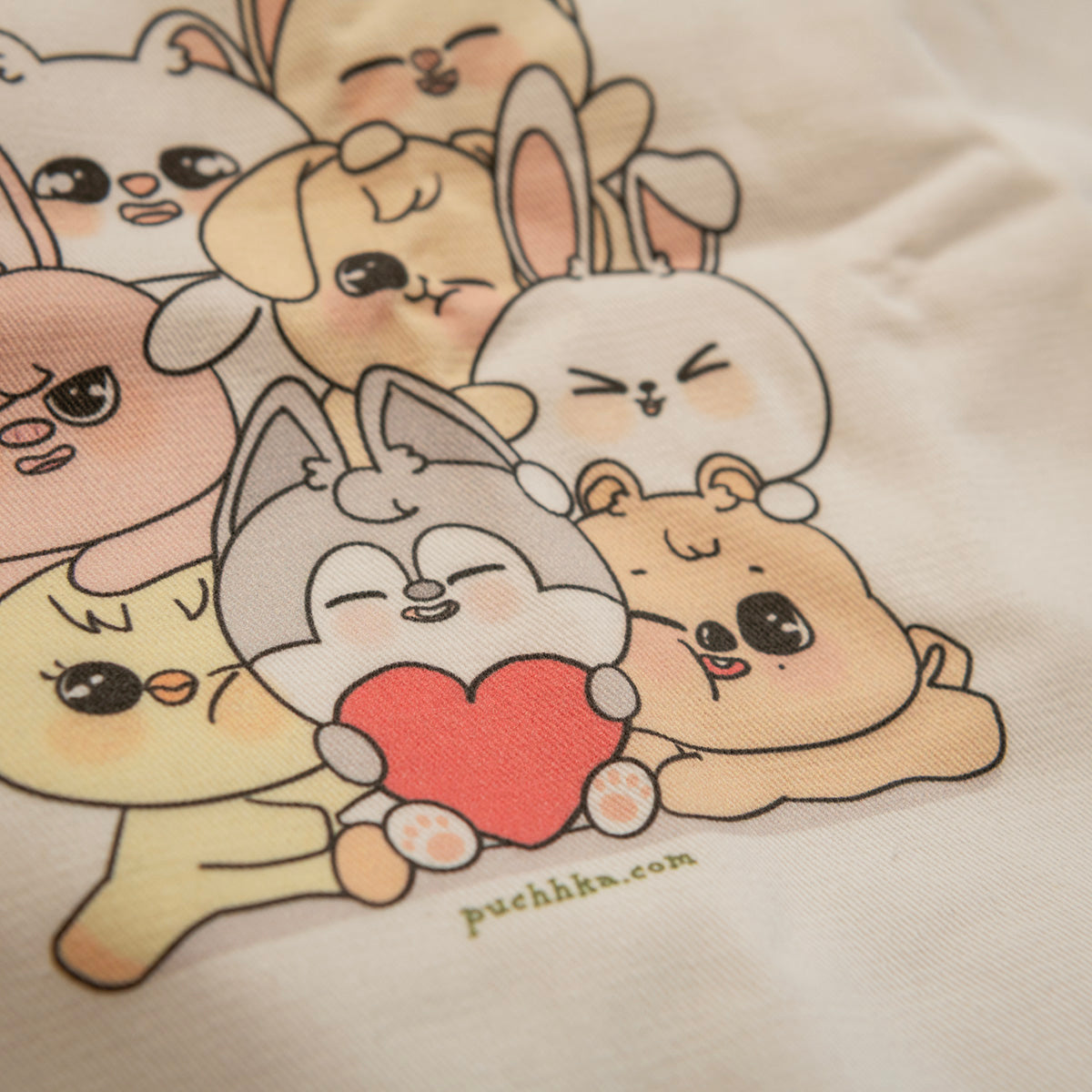 Close-up of a cream-colored fabric surface printed with a cute group of cartoon animals hugging and holding a red heart. The text 'puchhka.