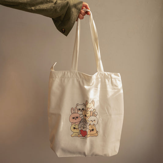 An off-white canvas tote bag with a cute cartoon rabbit and cat graphic, held by a hand wearing an olive green shirt against a plain beige wall.