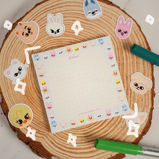 A square grid notepad with skzoo face border sits on a round wood coaster, surrounded by matching sticker cutouts and two colored markers.