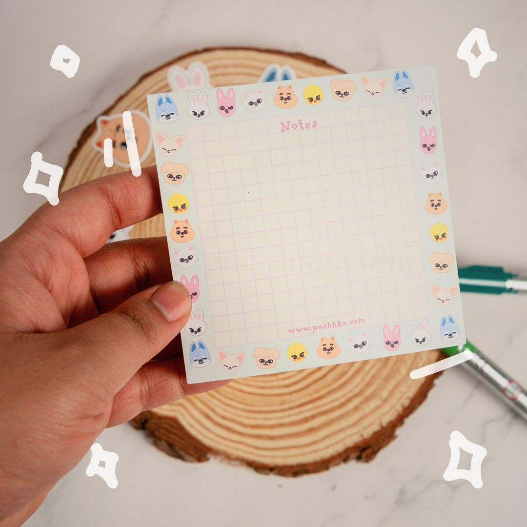 A hand holds a square grid notepad with a cute pastel border of skzoo. The notepad is photographed over a wood coaster with pens in the background.