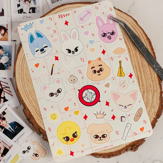 A sheet of cute skzoo stickers with expressive faces, hearts, stars, and small accessories, placed on a round wood slice with tweezers and printed photos nearby.