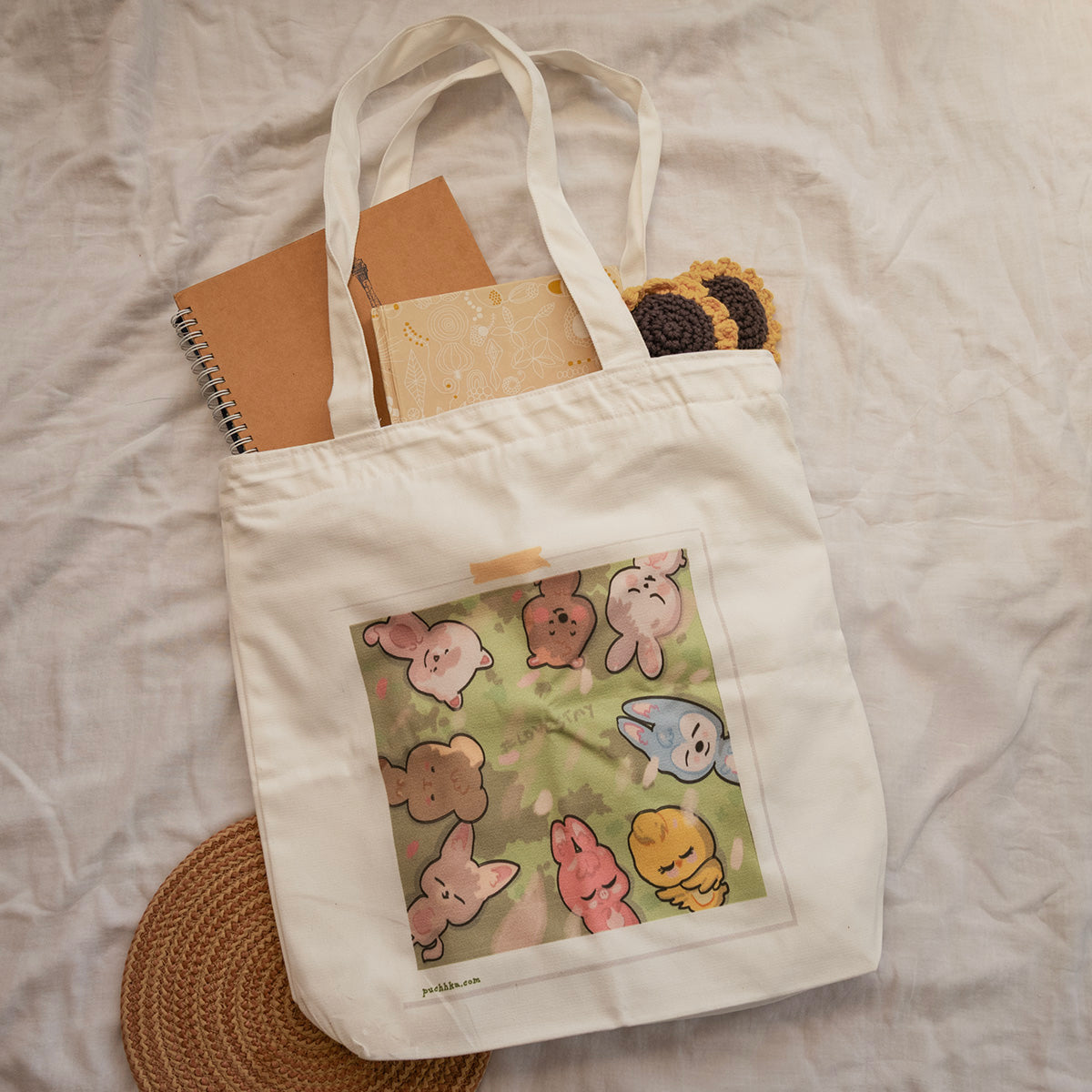 A cream-colored canvas tote bag with skzoo print on the front pocket, styled with notebooks and a crocheted coaster on a white fabric background.