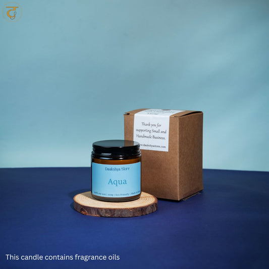 A small amber glass candle jar with a black lid and a light blue label reading 'Aqua' sits on a round wood slice coaster beside a brown kraft gift box. The scene is set on a dark blue surface against a soft blue backdrop, with a small text note at the bottom.