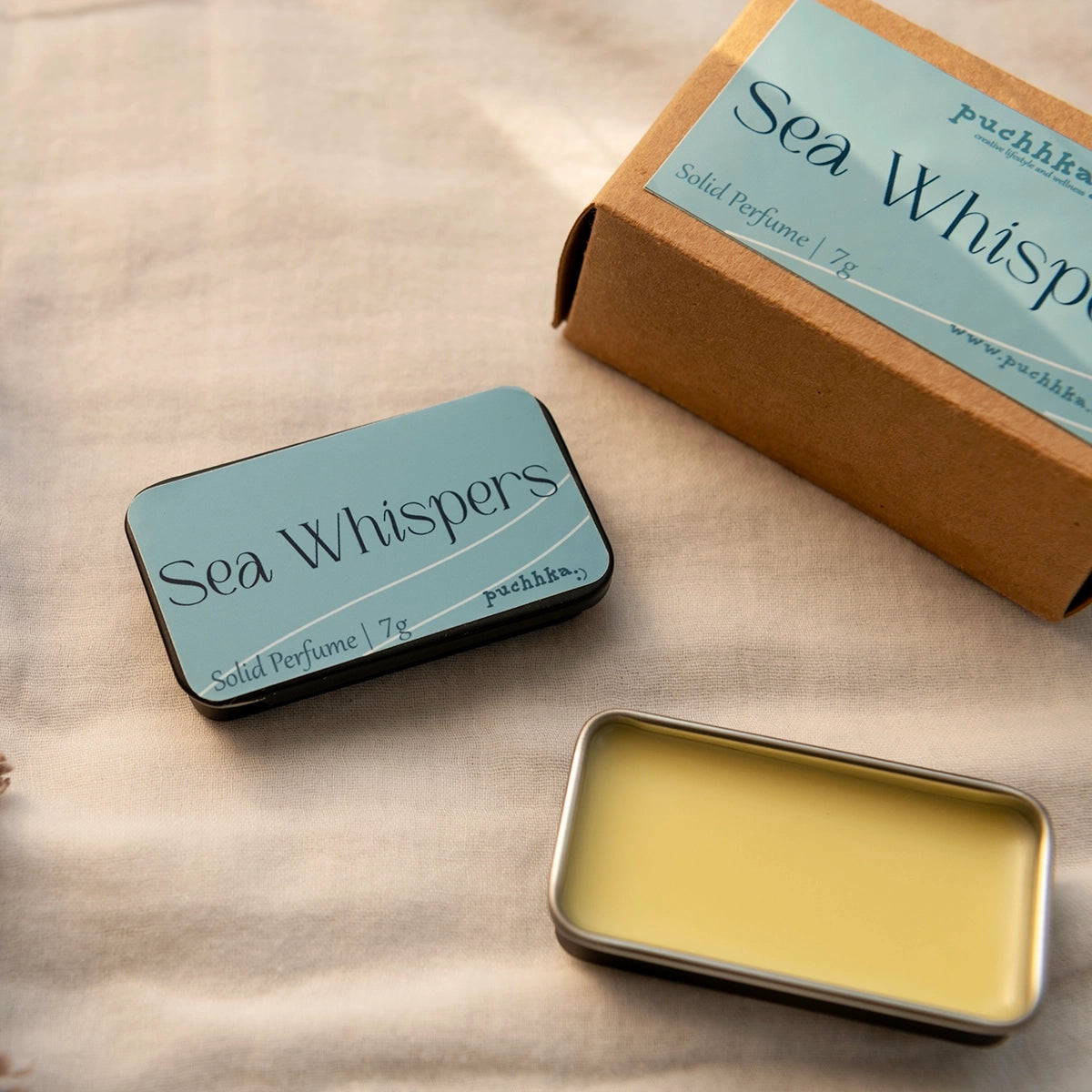 A tin of Sea Whispers solid perfume shown open with pale yellow scented wax inside, alongside its kraft paper box and closed tin with a light blue label, on a beige fabric background.