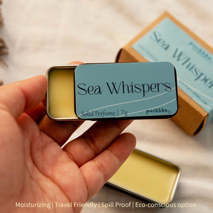 A hand holds an open silver tin of Sea Whispers solid perfume, showing the pale yellow balm inside and the light blue label. Another tin and its kraft paper box are blurred in the background on a soft white fabric surface.