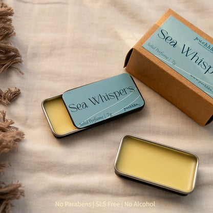Two small rectangular metal tins of Sea Whispers solid perfume with pale yellow wax, shown beside a kraft paper box on a beige fabric surface. 
