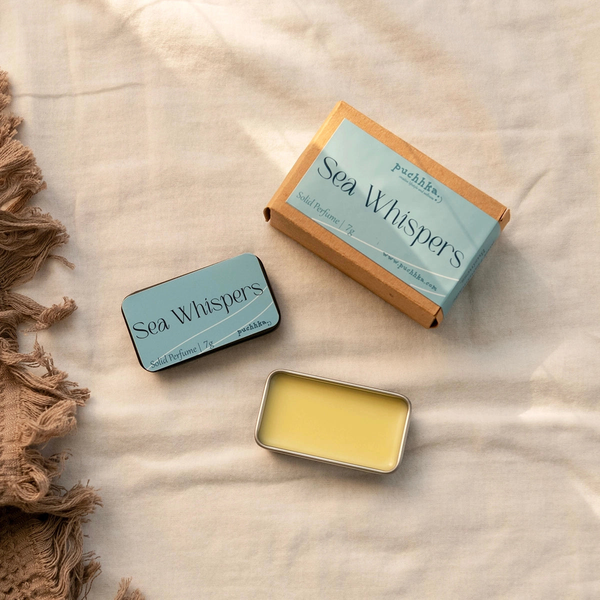 A flat-lay product photo of 'Sea Whispers' solid perfume by Puchhka. The image shows the light blue and kraft brown outer box, the matching square tin with the product name, and an open square tin displaying the pale yellow balm.