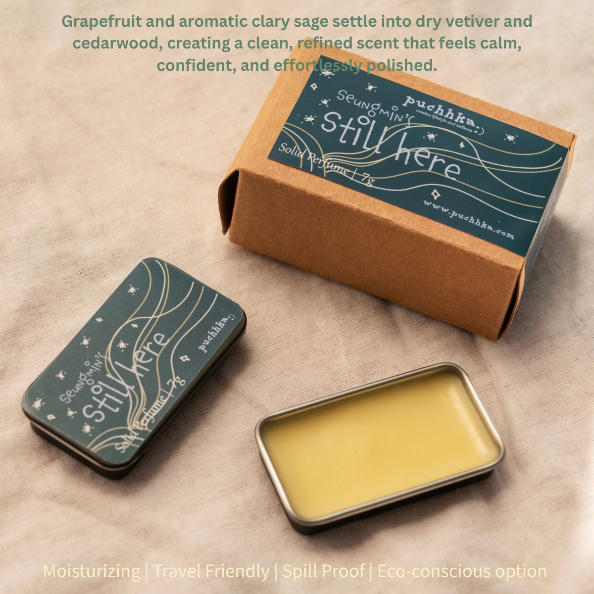 A small rectangular metal tin of pale yellow balm sits beside a closed kraft box and a matching closed tin, both printed with the scent name 'Seungmin's Still Here' and the brand 'Puchhka'. The scene is styled on a beige fabric background with soft text at the top and bottom describing the scent and benefits.