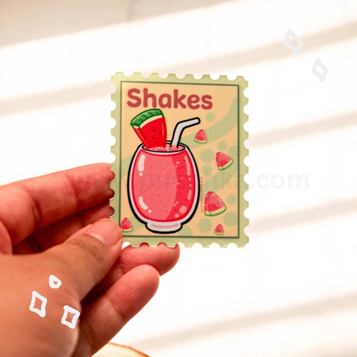 A hand holds a square sticker with a watermelon shake illustration and the word 'Shakes' on a light, softly lit background.