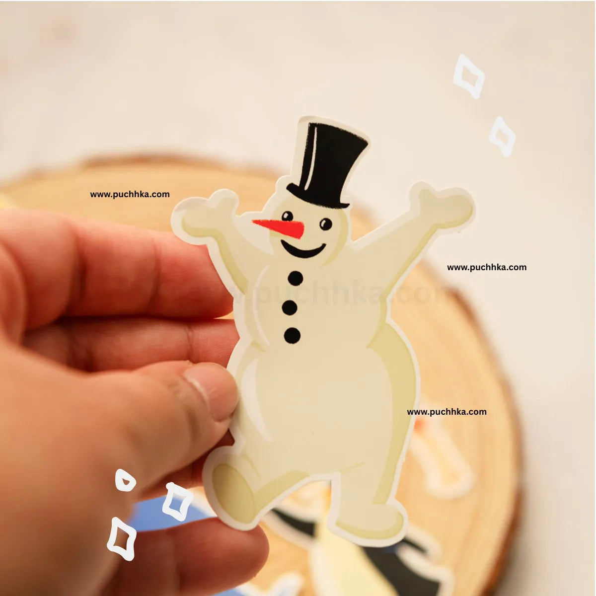 A hand holding a die-cut sticker of a smiling snowman wearing a black top hat and red carrot nose, with a blurred wooden surface in the background.
