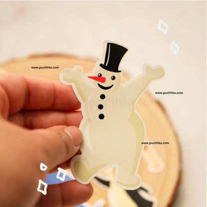 A hand holding a die-cut sticker of a smiling snowman wearing a black top hat and red carrot nose, with a blurred wooden surface in the background.