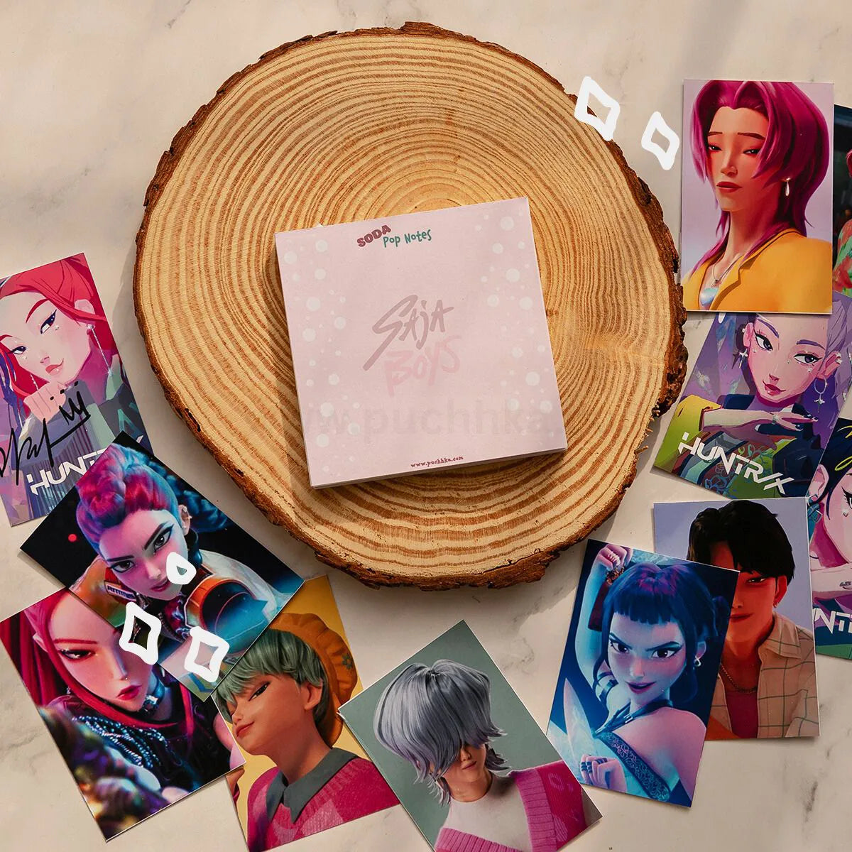 A square memopad 'Soda Pop Notes' sits centered on a round wood slice, surrounded by multiple illustrated character cards from Kpop Demon Hunters on a light marble-like surface.
