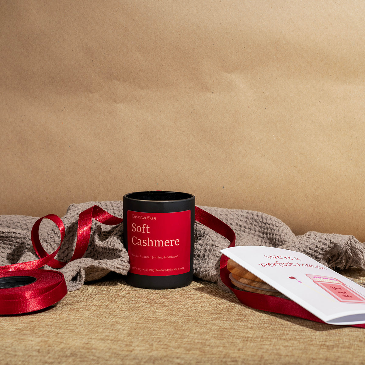 A amber glass jar candle with a red label reading 'Soft Cashmere' sits on a textured beige surface with a red ribbon, a folded knit scarf, and a small white card in the foreground, all against a brown kraft-paper backdrop.